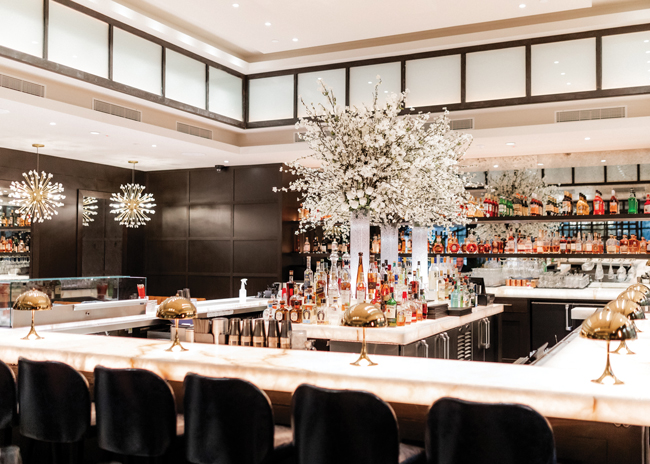 The bar at Truluck’s features an island with three large silk flower bouquets. The floral look is repeated  with nearby light fixtures.