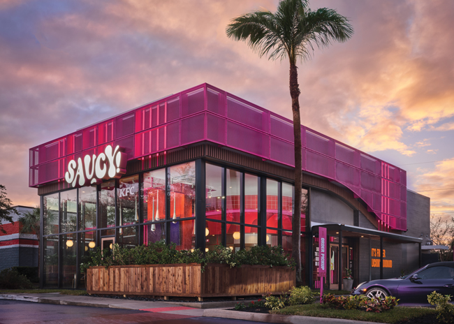 The building’s exterior features layers of material, such as signage on top of pink metal mesh, along with floor to ceiling windows.